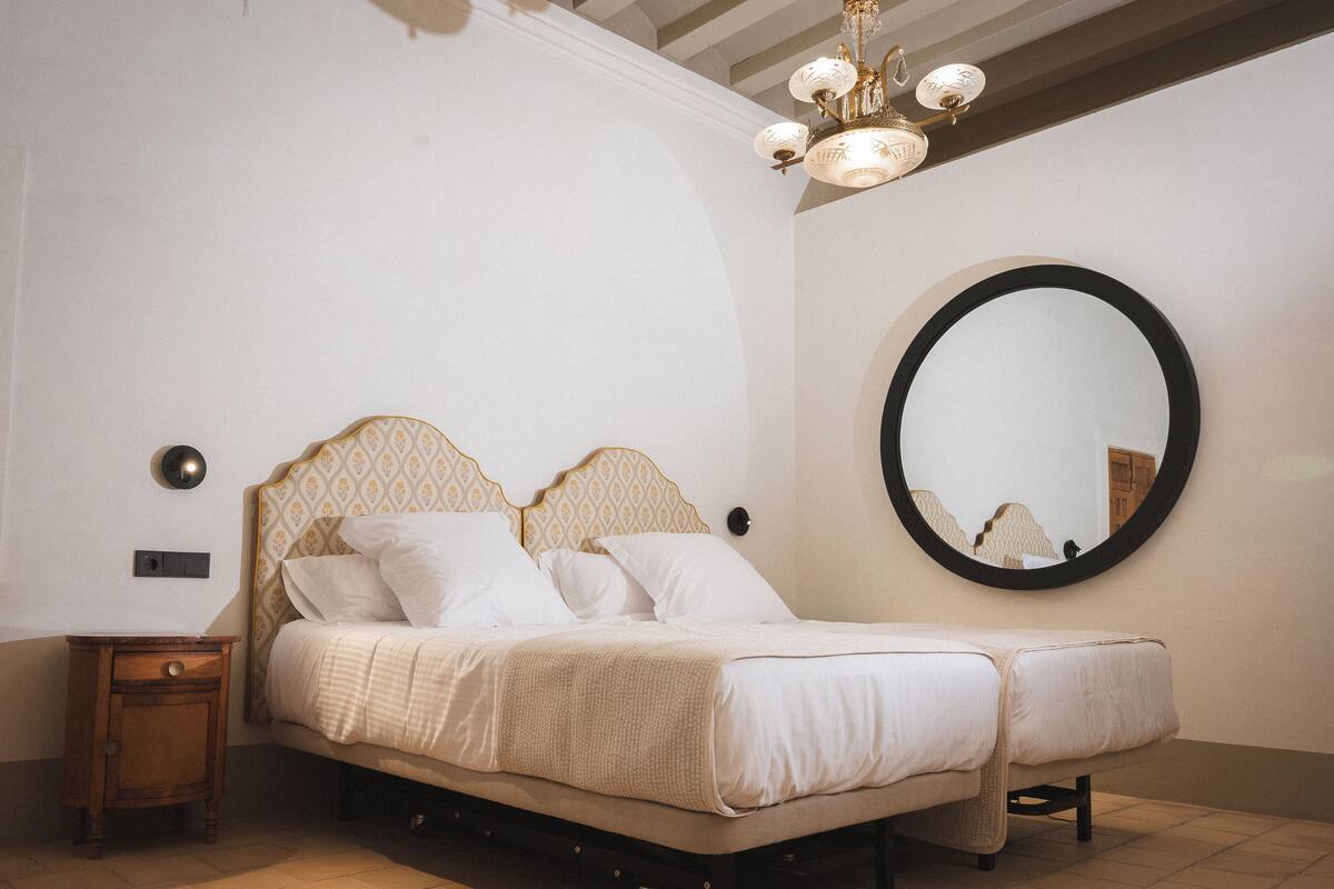 A bedroom with a bed, a nightstand, a round mirror, and a chandelier.