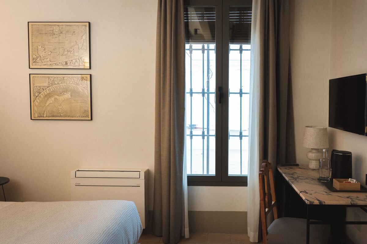 Habitación de hotel con una cama, un escritorio con lámpara, un televisor y dos mapas enmarcados en la pared.