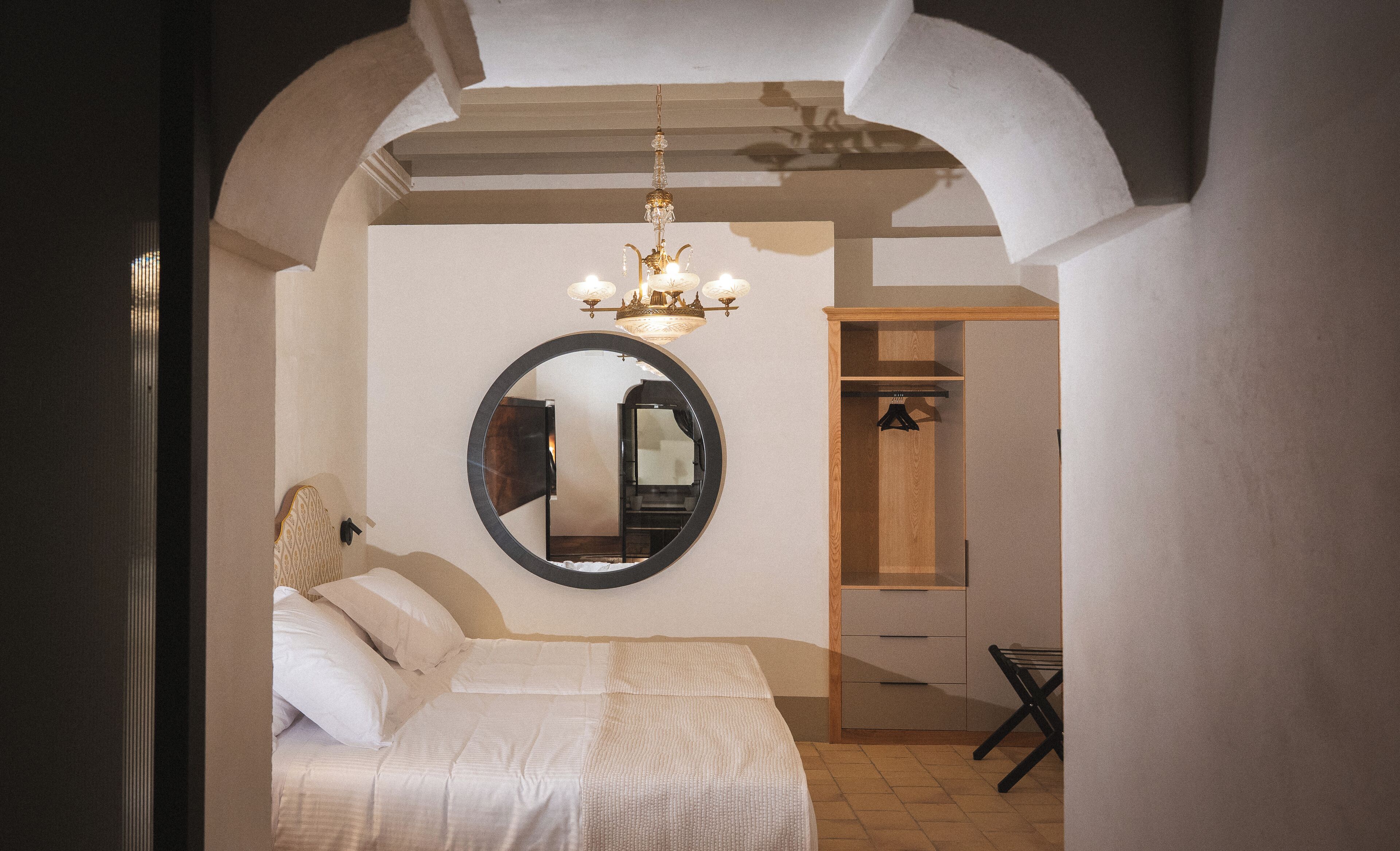 A bedroom with a large bed, a round mirror, a chandelier, and a wooden wardrobe.