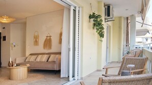 A spacious balcony with wicker chairs, a round wooden table, and a sliding glass door leading to a living area with a sofa and decorative wall hangings. - Modern Beach Retreat 2BR in Coastal Palaio Faliro (Paleo Faliro)