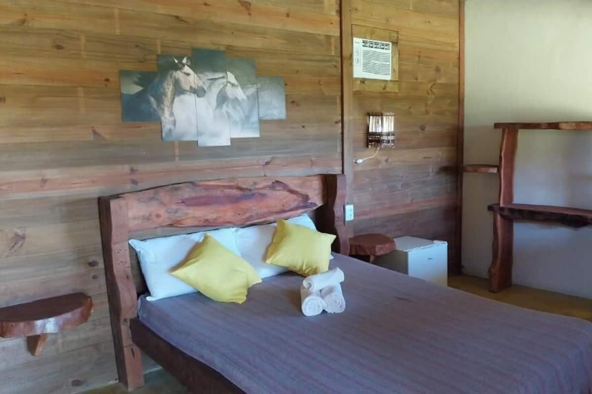 A wooden cabin room with a bed, two pillows, a wall-mounted light, and a wooden chair.