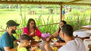 Uma família jantando em uma mesa de madeira ao ar livre, cercada por vegetação.