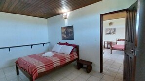 Quarto com cama, mesa de cabeceira, cadeira e porta que dá para outro cômodo.