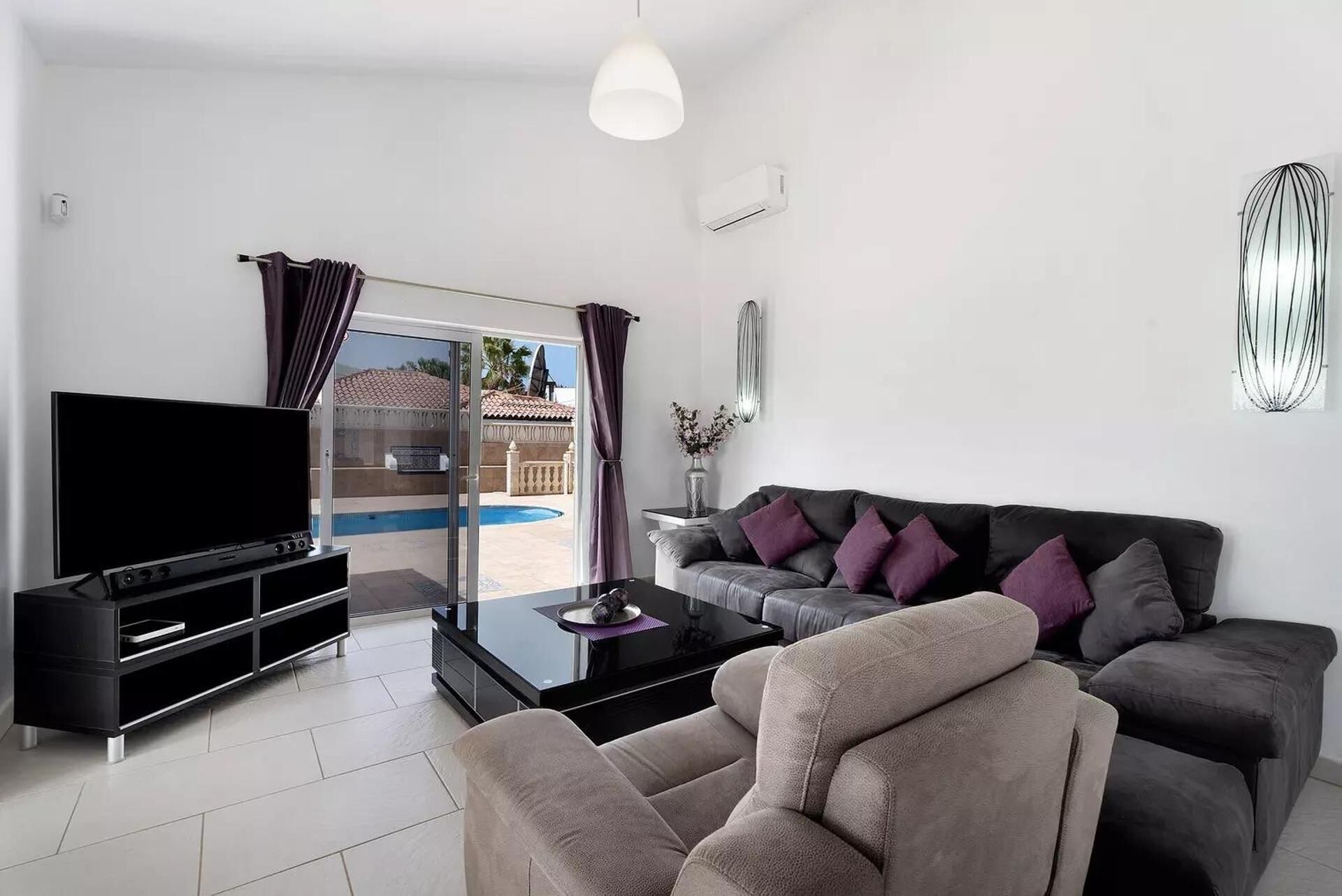 Un saló modern amb un televisor de pantalla plana, un sofà amb coixins, una taula de centre i una porta de vidre que dona a una zona de piscina exterior.