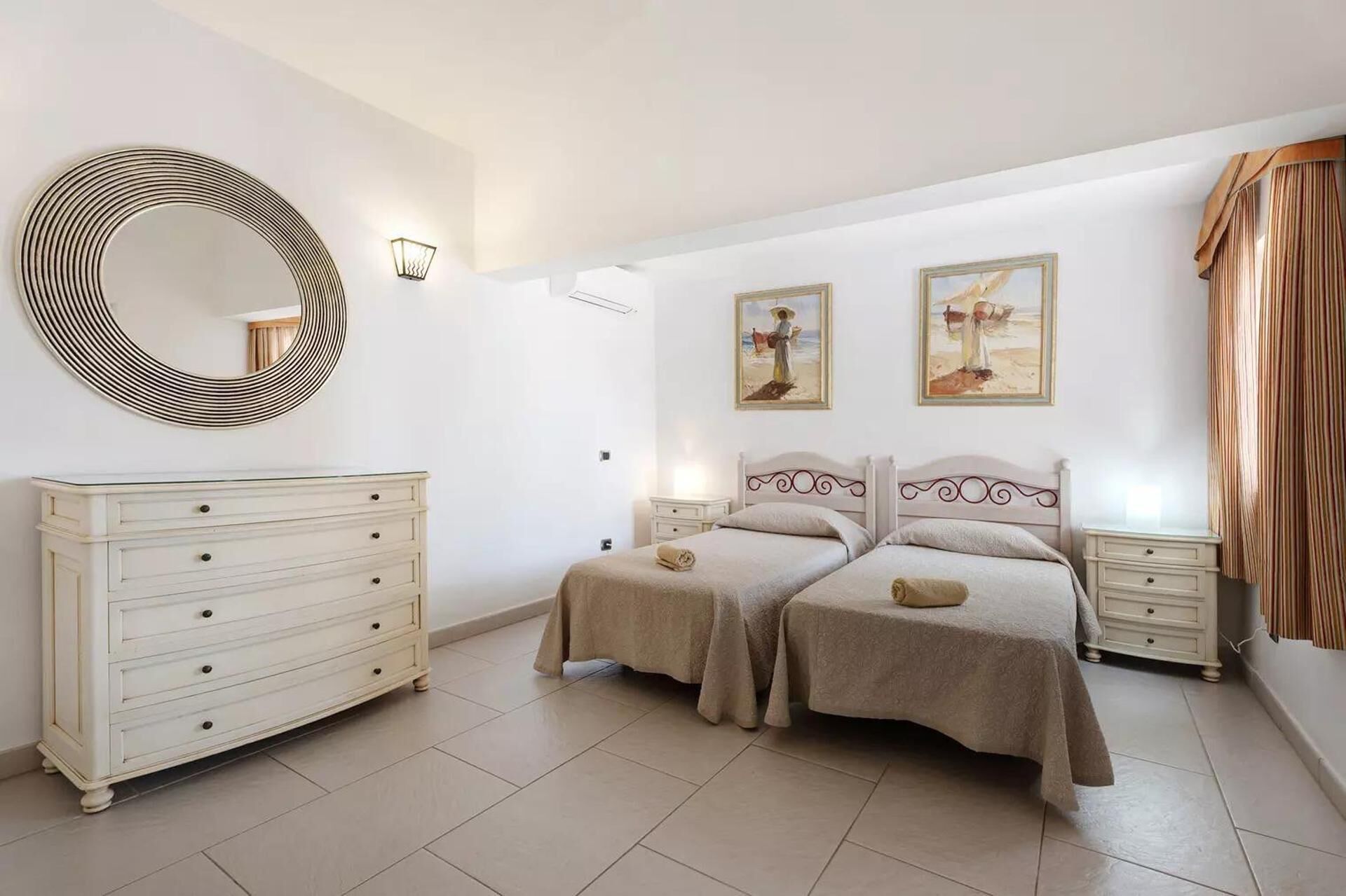 Un dormitorio con dos camas, un aparador, un espejo redondo y dos cuadros colgados en la pared.