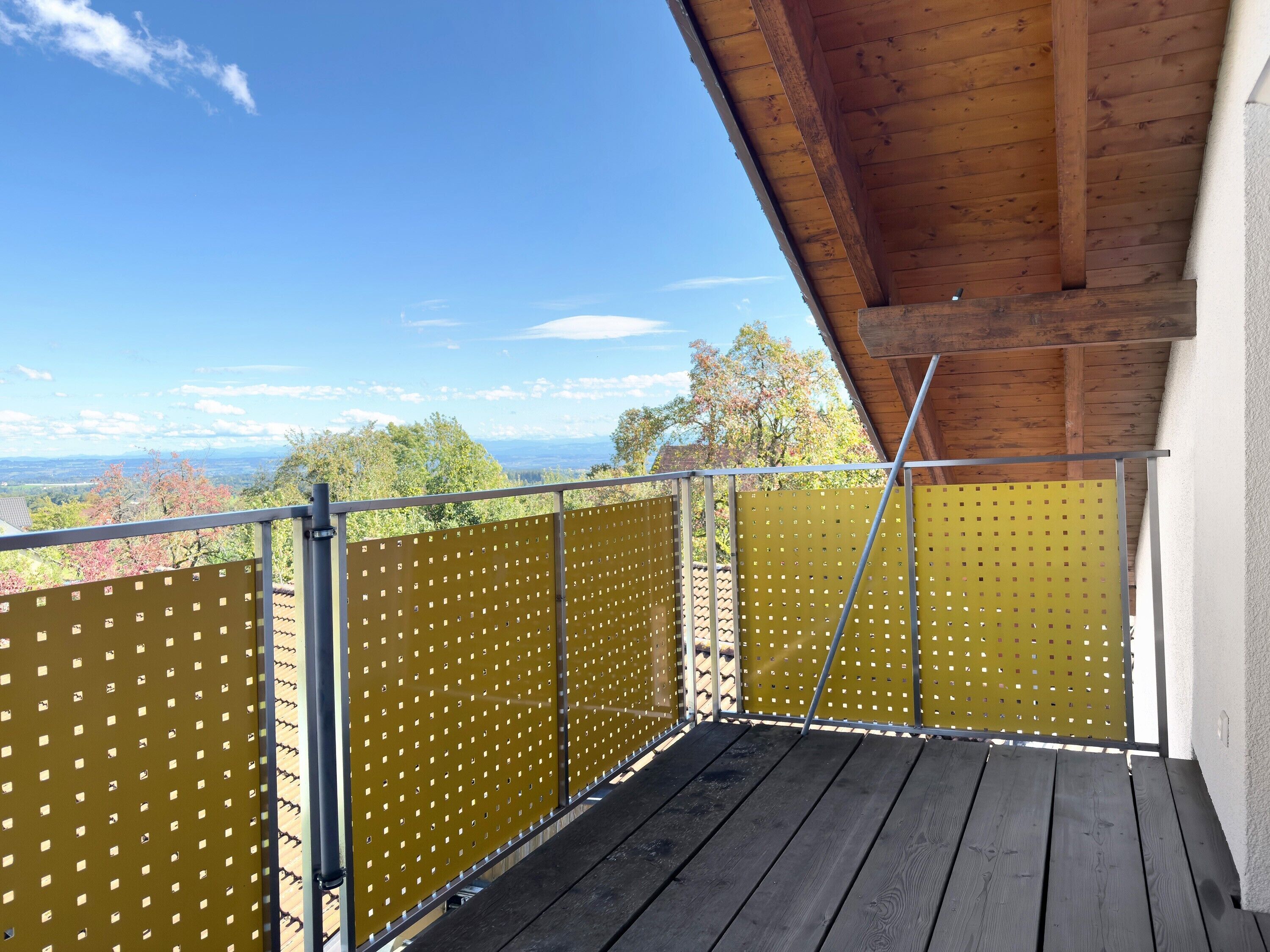 Un balcon doté d’un plancher en bois et de rampes en métal, offrant une vue panoramique sur un paysage verdoyant, avec des arbres et des montagnes à l’horizon.