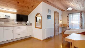 Een kamer met houten plafond, witte muren, een kitchenette met televisie, een bed en een raam met gordijnen.