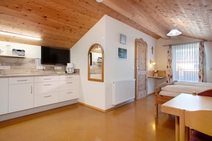 Une pièce avec un plafond en bois, des murs blancs, un coin cuisine avec une télévision, un lit et une fenêtre avec des rideaux.