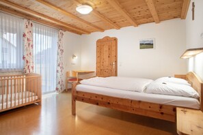 Un lit en bois avec une literie blanche, un berceau en bois, un bureau en bois et une armoire en bois dans une pièce dont le plafond est orné de poutres en bois et où un tableau est accroché au mur.