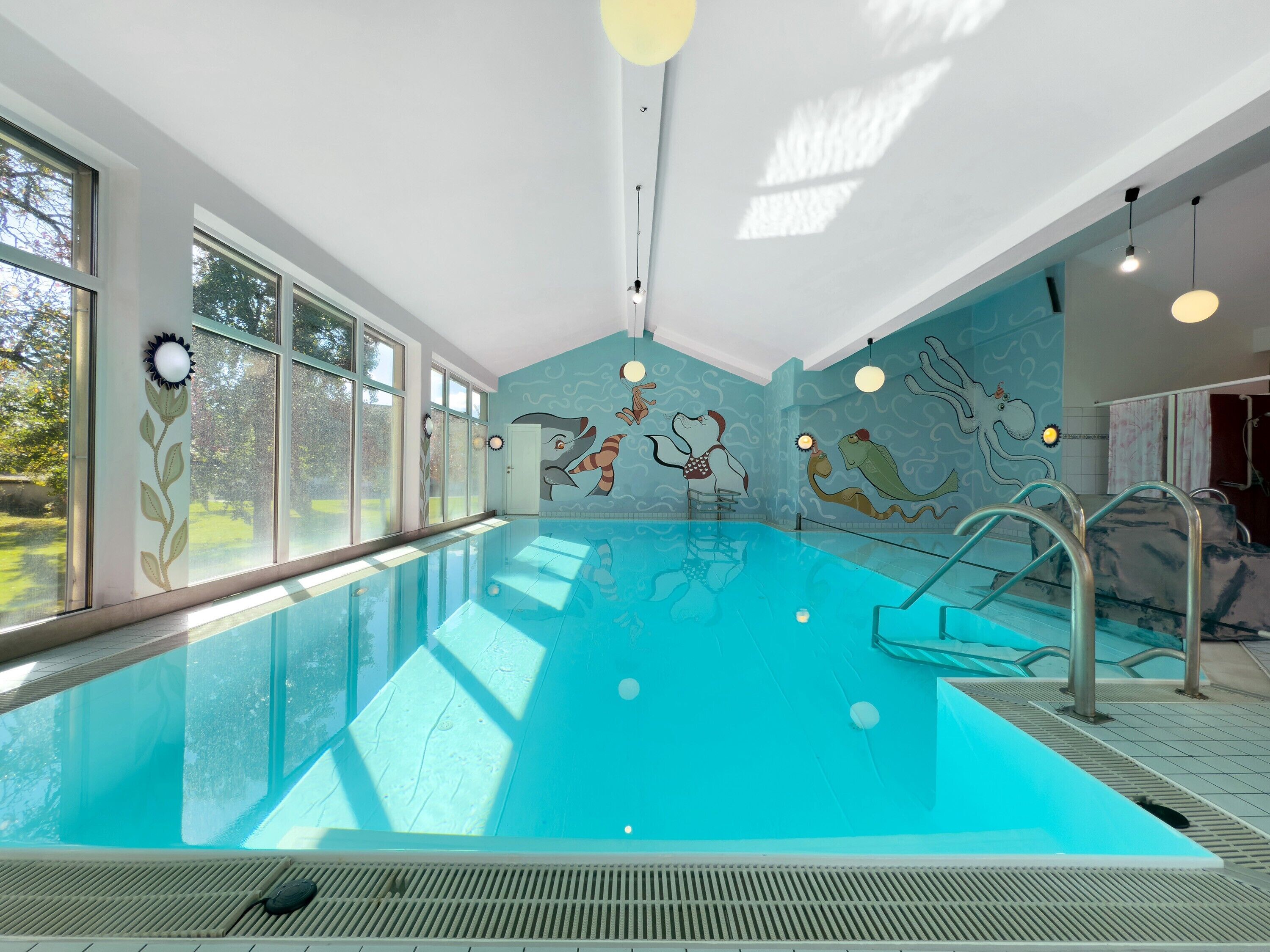 An indoor swimming pool with clear glass walls, a mural with aquatic themes, and a sunlit ceiling.