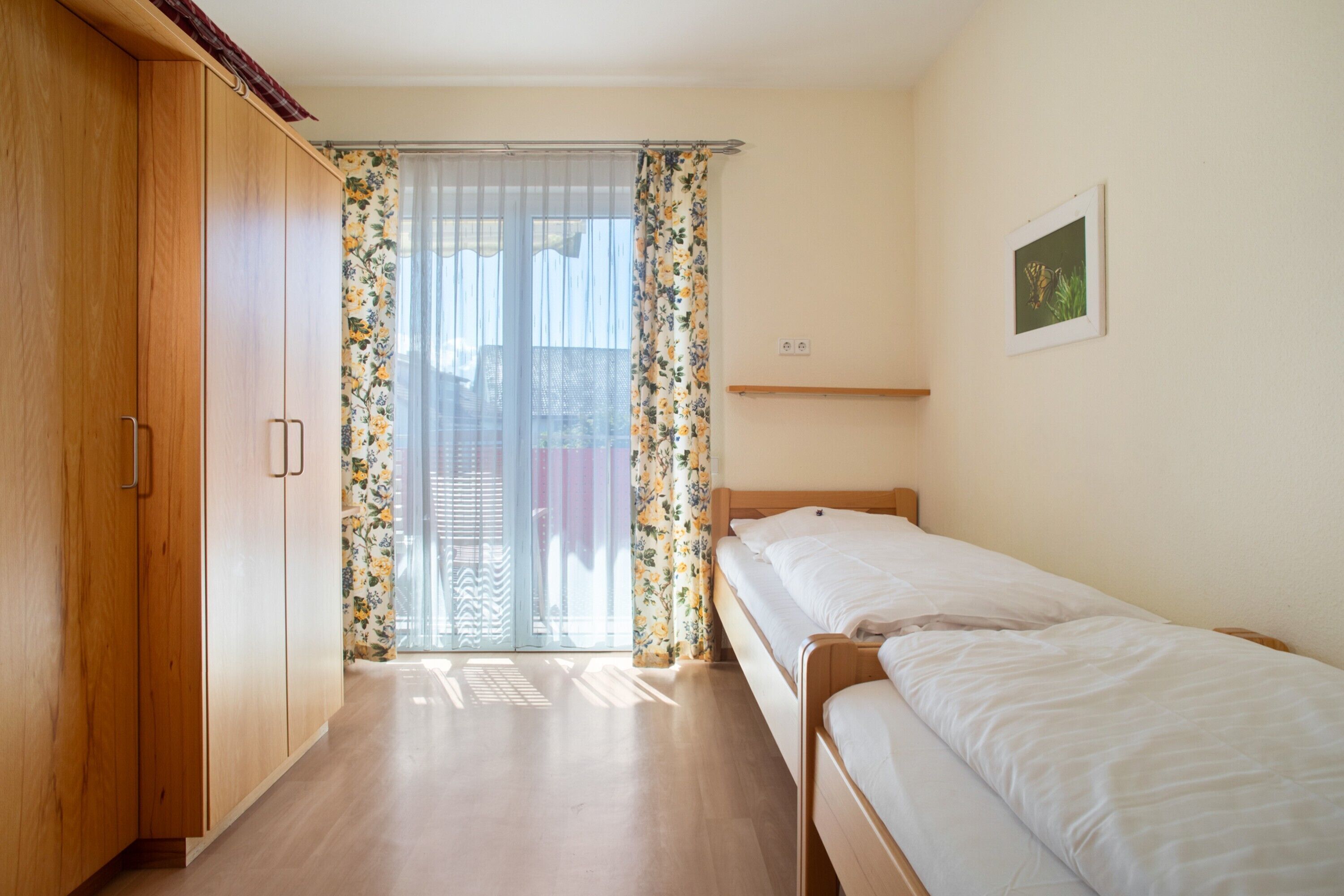 Ein Schlafzimmer mit einem Holzschrank, einem Einzelbett, einem Fenster mit blumigen Vorhängen und einer kleinen Ablagefläche.