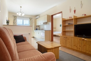 A living room with a sofa, a wooden coffee table, a TV on a wooden stand, a kitchen in the background, and a window with curtains.