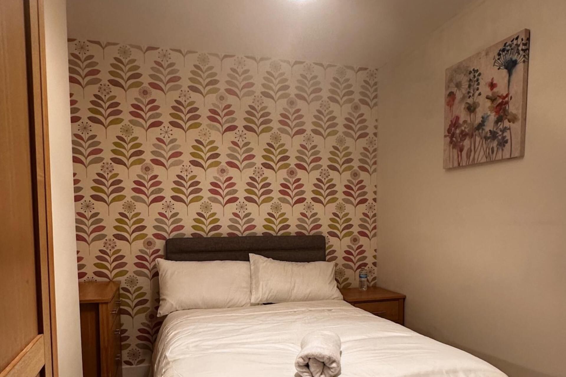 A bedroom with a floral wallpaper, a wooden headboard, a white bedspread, and a painting on the wall.