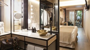 Une chambre d’hôtel moderne avec un grand miroir, une coiffeuse avec éclairage, un bol doré, un lit recouvert d’une couverture à motifs et une vue sur le balcon.