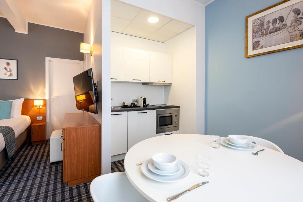A compact room with a bed, a small bedside table, a desk with a chair, a kitchenette, and a dining area with a table set for two.