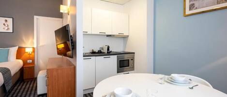 A compact room with a bed, a small bedside table, a desk with a chair, a kitchenette, and a dining area with a table set for two.
