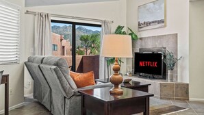 Un salon avec un canapé gris, une table basse en bois, un téléviseur affichant le logo « NETFLIX », une cheminée et une vue sur les montagnes par une grande fenêtre.
