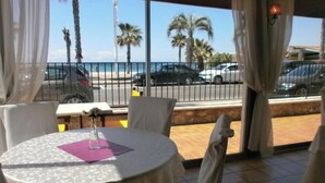 Un comedor con una mesa dispuesta para dos personas, un florero con flores y vistas a la calle y palmeras.