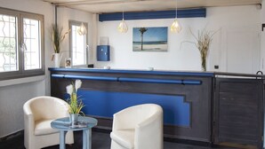 A reception area with a blue counter, two white armchairs, a small round table with a vase, and a beach-themed picture on the wall.