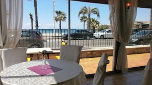 Un comedor con una mesa dispuesta para dos personas, un florero con flores y vistas a la calle y palmeras.