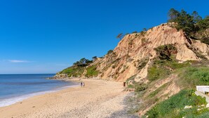 A beach with a rocky cliff, clear blue sky, and a few people walking along the shore.