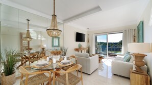 A modern living room with a glass dining table, wicker chairs, a sofa, and a TV mounted on the wall.