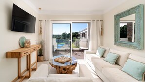 A modern living room with a flat-screen TV, a wooden console table, a glass coffee table, a beige sofa with teal cushions, and a large mirror.