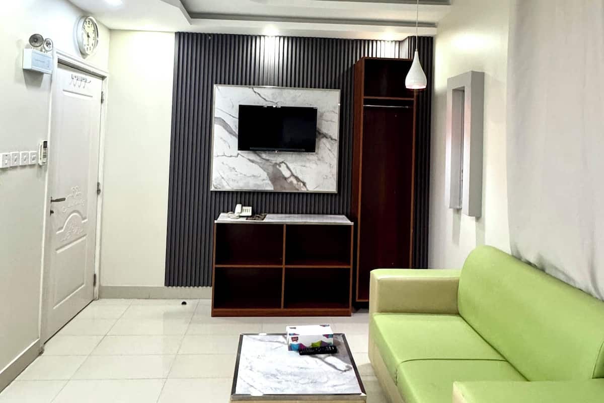 A modern living room with a flat-screen TV mounted on a marble wall, a green sofa, a beige ottoman, and a tiled floor.