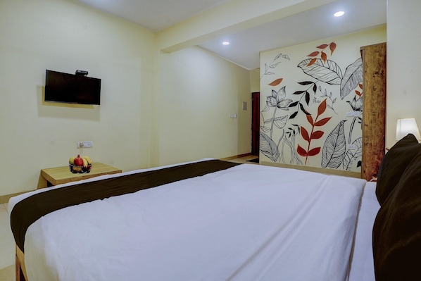 A hotel room with a bed, a television mounted on the wall, a small table with a fruit bowl, and a wall with a floral mural.