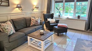 Un salon comprenant un canapé, un fauteuil et une table basse. On y trouve deux lampes fixées au mur, une fenêtre avec des rideaux et un radiateur.