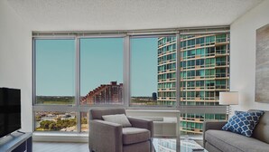 Ein modernes Wohnzimmer mit Sofa, Sessel und Glastisch, großen Fenstern mit Blick auf die Stadt und einem Flachbildfernseher.