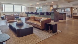 Eine moderne Hotelhalle mit einer Couch, Sesseln, einem Couchtisch, einer Bar und einem großen Fenster, das natürliches Licht hereinlässt.