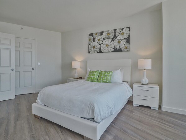 A neatly arranged bedroom with a bed, two bedside tables with lamps, a white door, and a floral artwork on the wall.