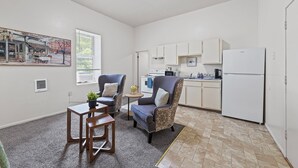 Un ambiente abitativo compatto con angolo cottura, una zona pranzo con due sedie e un tavolino, e un'ampia finestra che permette l'ingresso di luce naturale.
