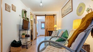 Ein gemütliches Wohnzimmer mit einer Couch, einem Sessel, einem Fernseher, einem Bücherregal und einer Schiebetür, die in einen anderen Raum führt.