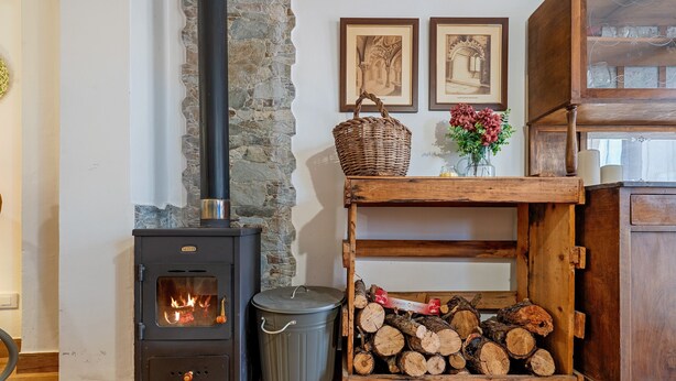 Ein Kaminofen, ein Mülleimer, ein Holzstoß, ein Korbwägelchen und zwei gerahmte Bilder an der Wand.