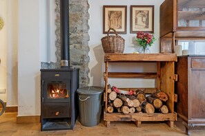 A wood-burning stove, a trash bin, a stack of firewood, a wicker basket, and two framed pictures on the wall. - L'escambell Grau del Llop – Rural Tourism Apartment (Ulldemolins)