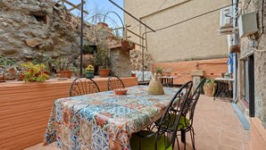 Un patio al aire libre con una mesa cubierta por un mantel estampado, rodeada de sillas, plantas en macetas y un muro de piedra.