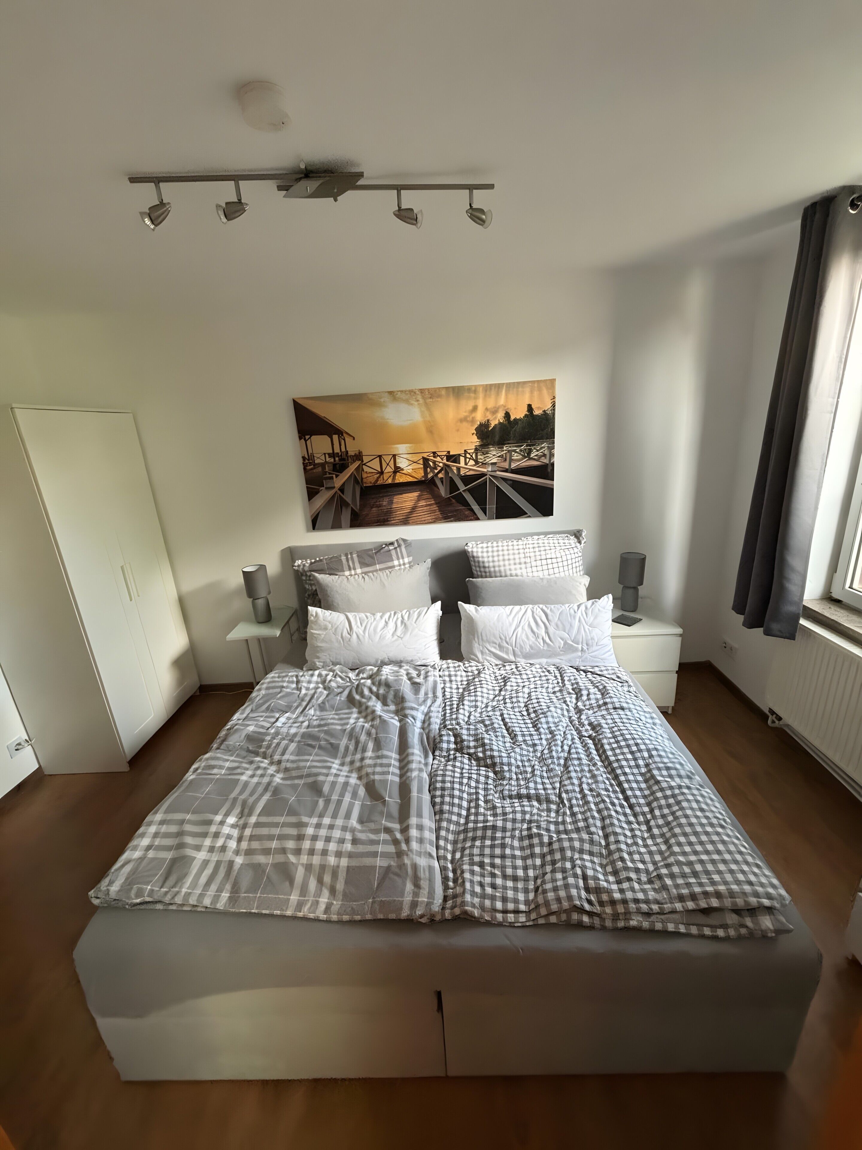 A bedroom with a bed, white nightstands, a painting, a window, and a door.