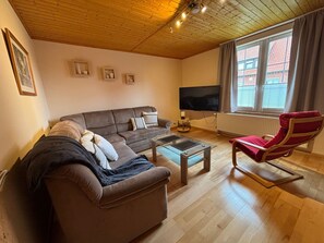 A living room with a sofa, a red chair, a coffee table, a TV, and a window with blinds. - Heiderast Wipperau with Private Terrace, Garden, and Wi-Fi (Oetzen)