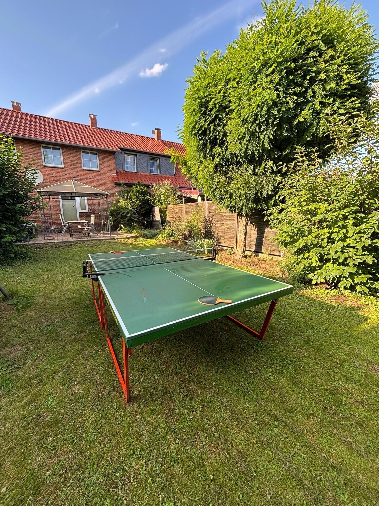 Ein grüner Tischtennis-Tisch in einem Garten mit einem Ziegelhaus und einem großen Baum.