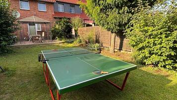Ein grüner Tischtennis-Tisch in einem Garten mit einem Ziegelhaus und einem großen Baum.