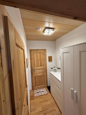 A hallway with wooden walls, a wooden door, a white dresser, and a small bedside table with a vase of flowers. - Heiderast Wipperau with Private Terrace, Garden, and Wi-Fi (Oetzen)