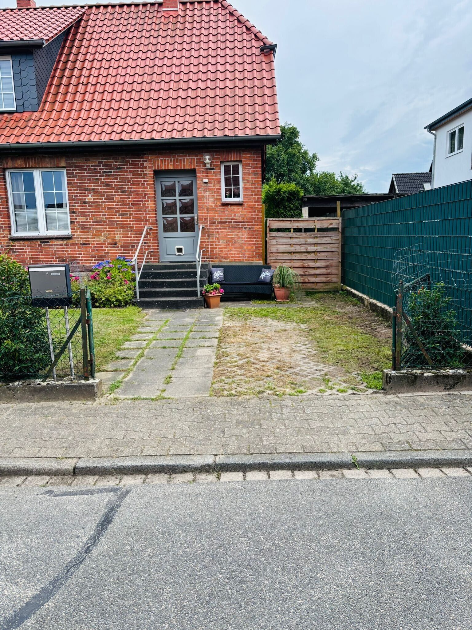 Une maison en briques avec un toit recouvert de tuiles rouges, une porte d’entrée et un petit jardin devant, agrémenté d’un banc et de plantes en pot.