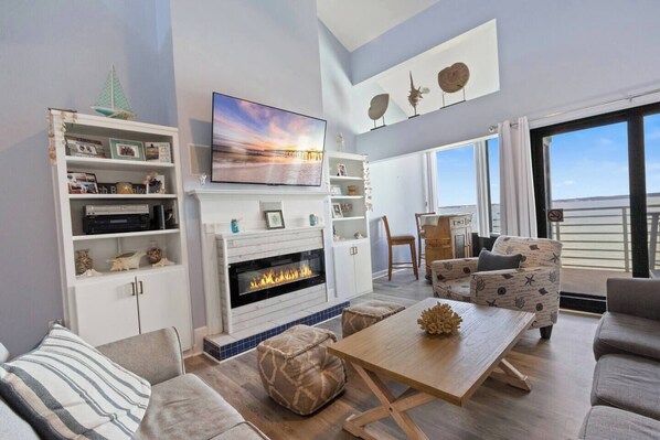Una sala de estar moderna con chimenea, asientos cómodos, un televisor montado en la pared y vistas al mar a través de amplios ventanales.
