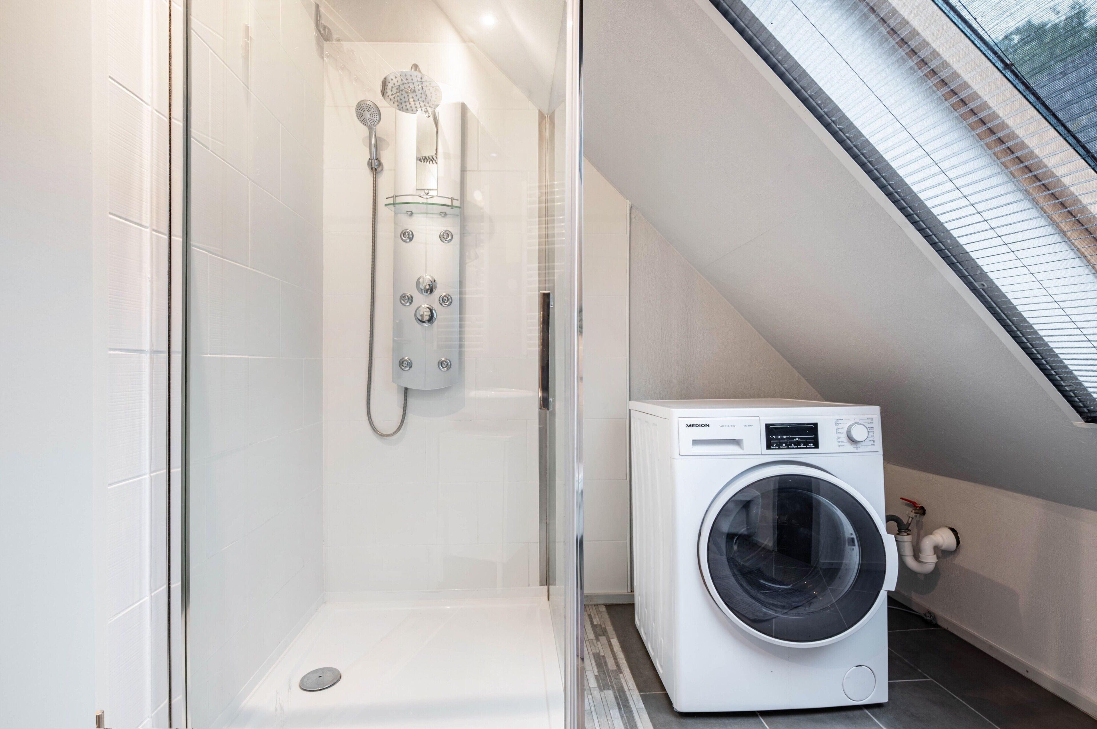 Un baño moderno con cabina de ducha de vidrio, lavadora integrada y techo inclinado con tragaluces.