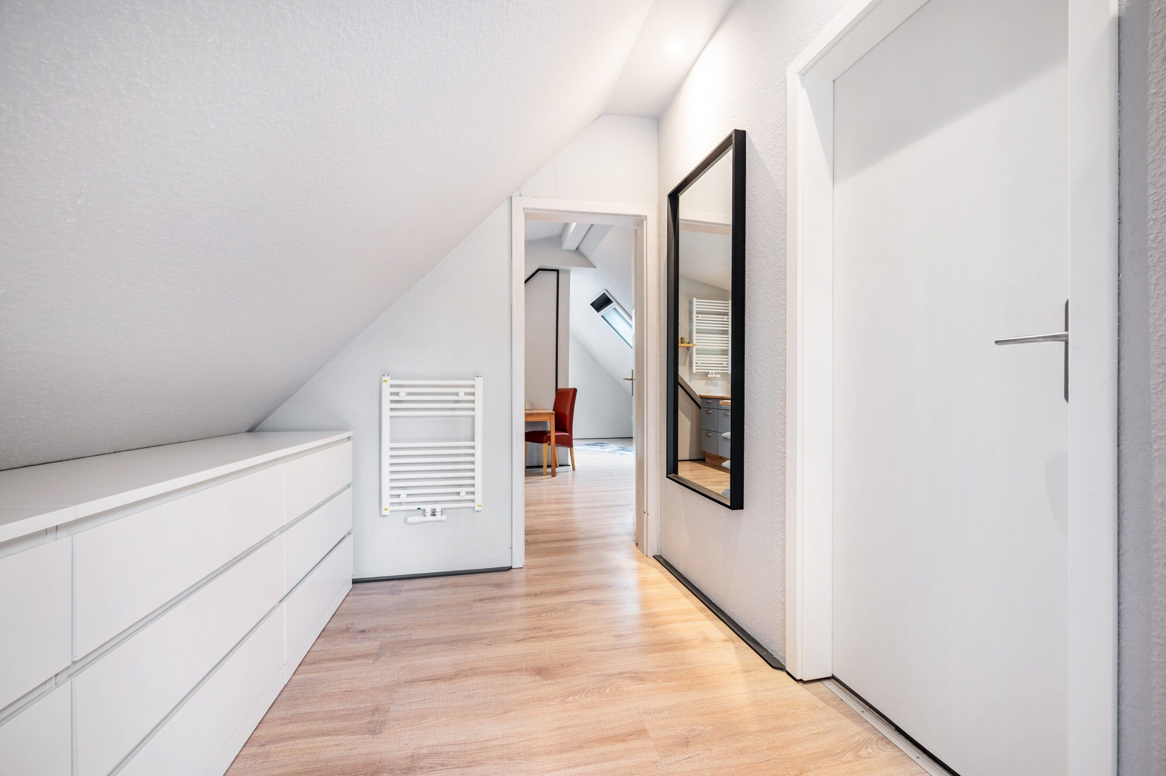 Un corridoio stretto con soffitto spiovente, una parete bianca con uno specchio e un pavimento in legno.
