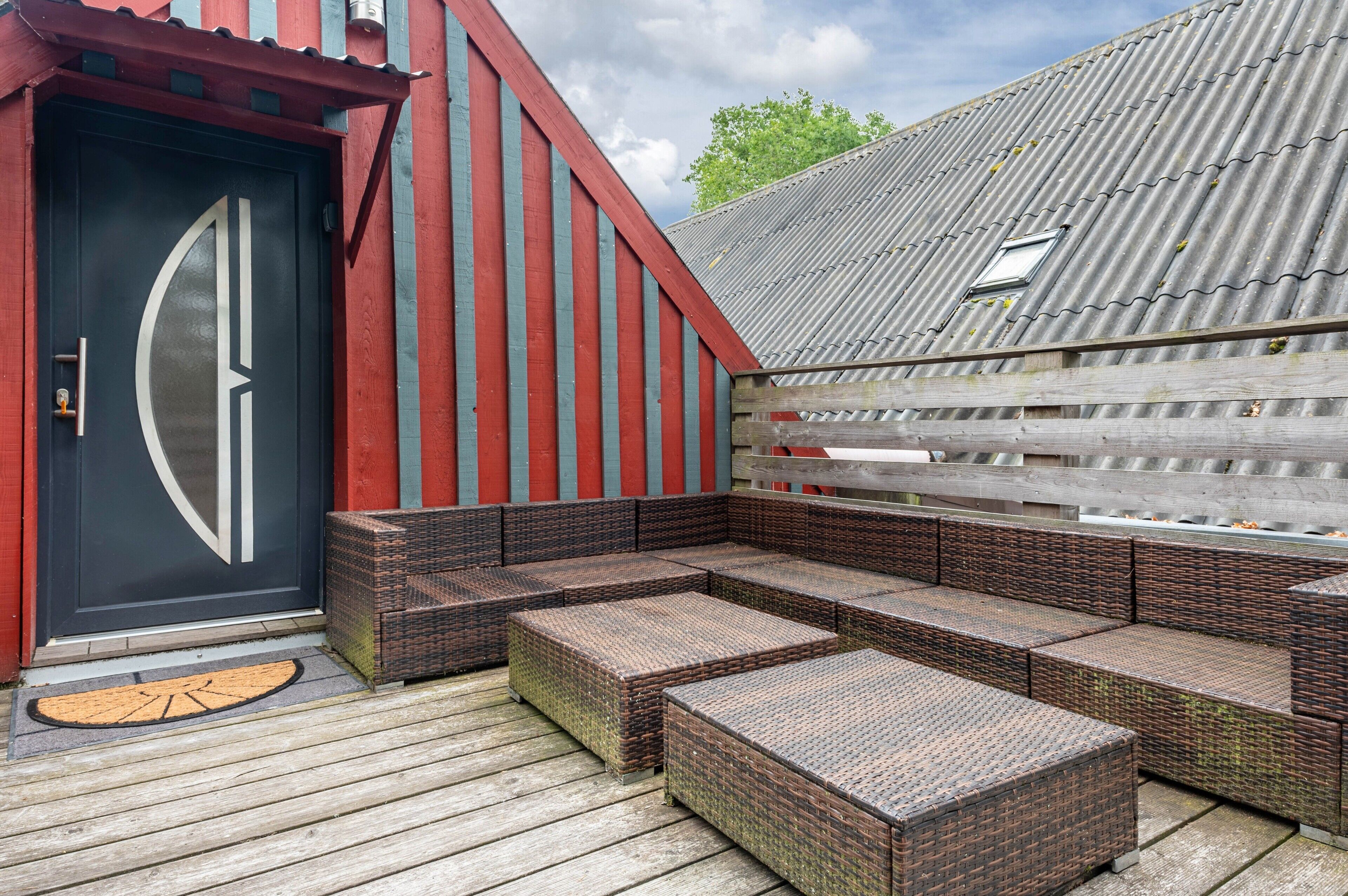 A covered outdoor seating area with wicker furniture, a black door with a geometric design, and a corrugated metal roof.