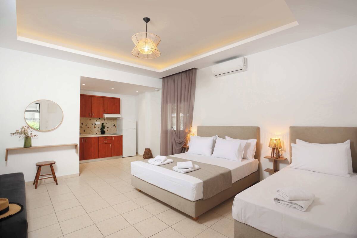 Habitación de hotel con cama doble, mesita de noche con lámpara, un taburete pequeño y vista al área de la cocina.