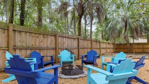 Un patio trasero con un cerco de madera, un pozo de fuego y varios sillones Adirondack azules dispuestos alrededor.